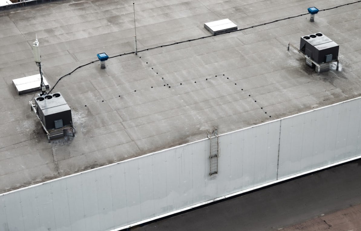 Aerial view of a large commercial flat roof with rooftop HVAC units, vents, skylights, and visible weathering on the roof surface.
