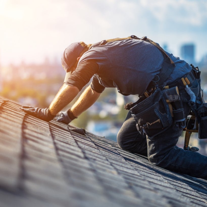 Roofer installing asphalt shingles on a residential roof while wearing a tool belt and safety equipment.