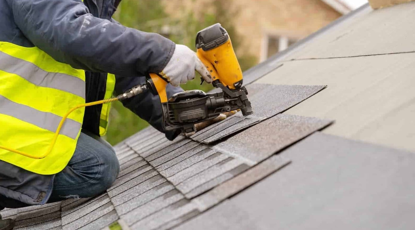 A worker wearing a high-visibility safety vest uses a yellow pneumatic nail gun to fasten asphalt shingles onto a roof.