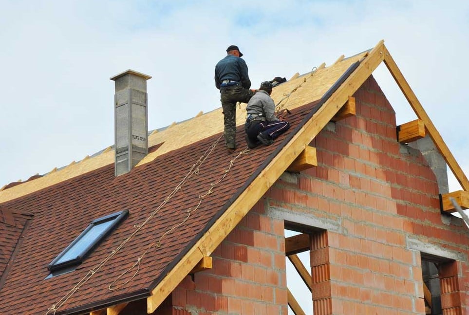 Two construction workers secure safety ropes as they install reddish-brown shingles on a newly built brick house with a tall chimney.