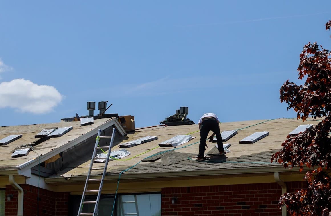 Roofer installing asphalt shingles on a residential roof with roofing materials, exposed decking, and a ladder visible.