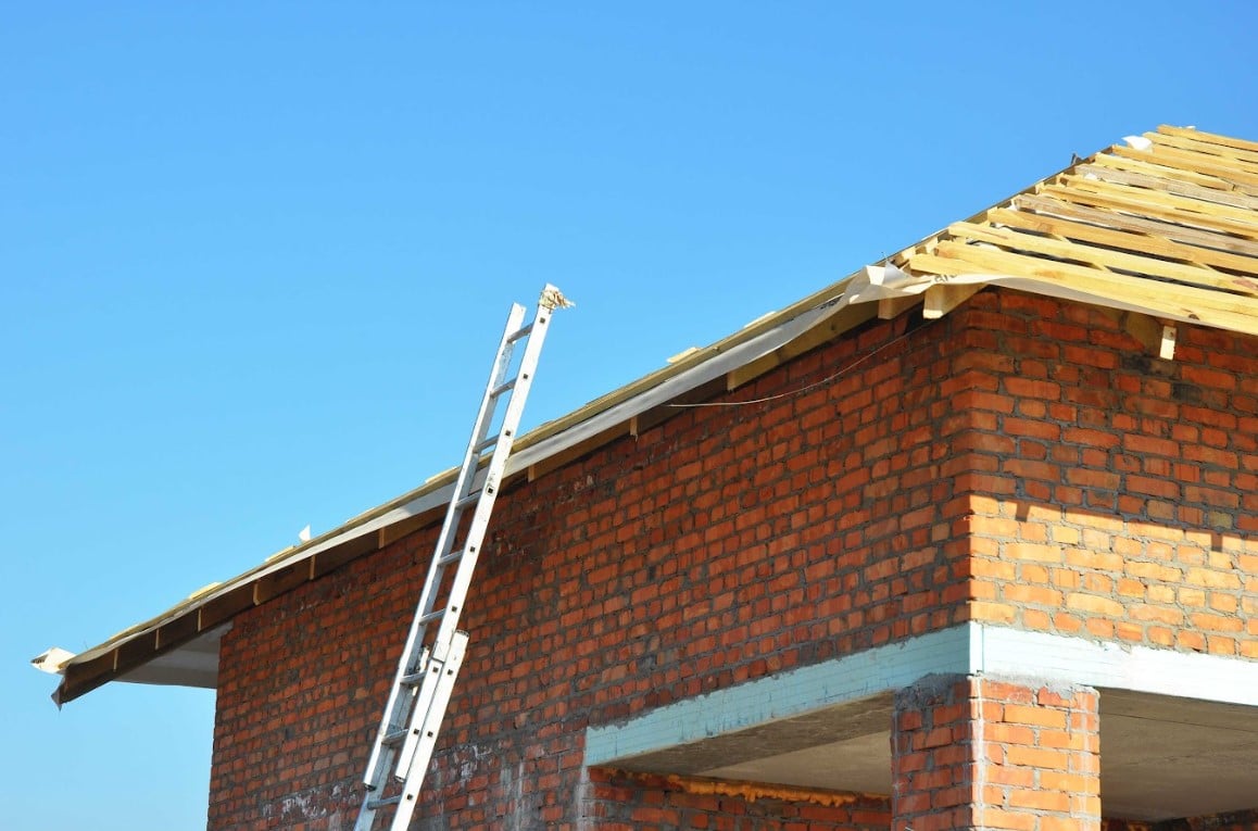 Residential brick building under construction with exposed roof framing and a ladder leaning against the exterior.