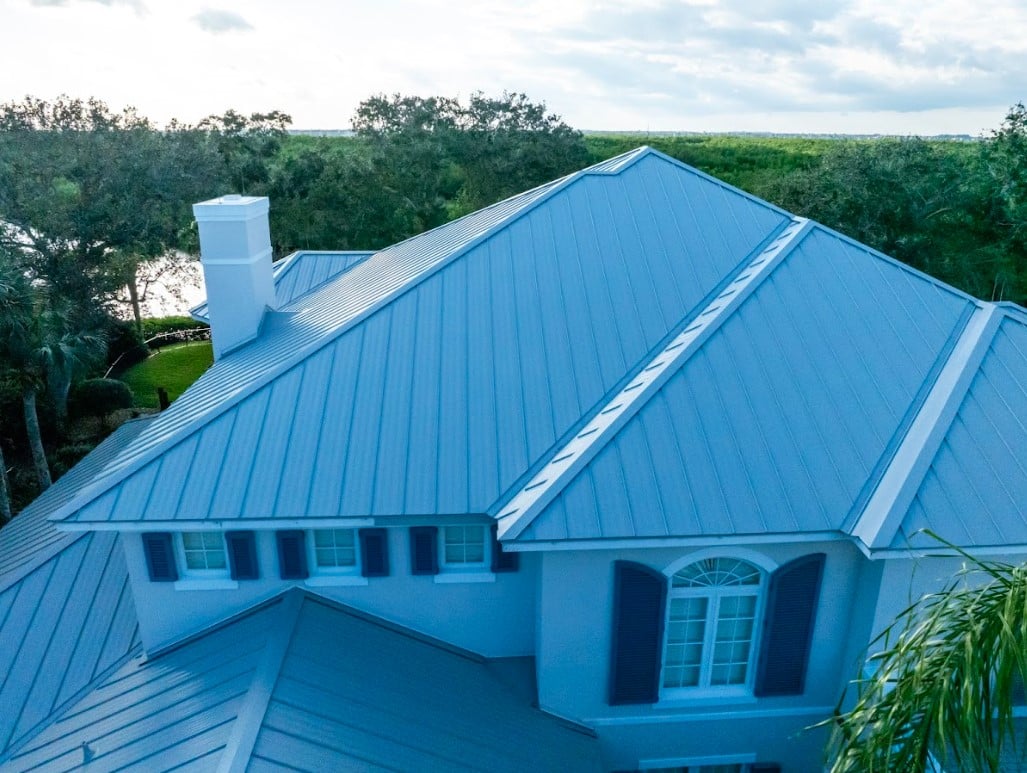 Aerial view of a Florida home with a light standing seam metal roof, white chimney, and surrounding trees.