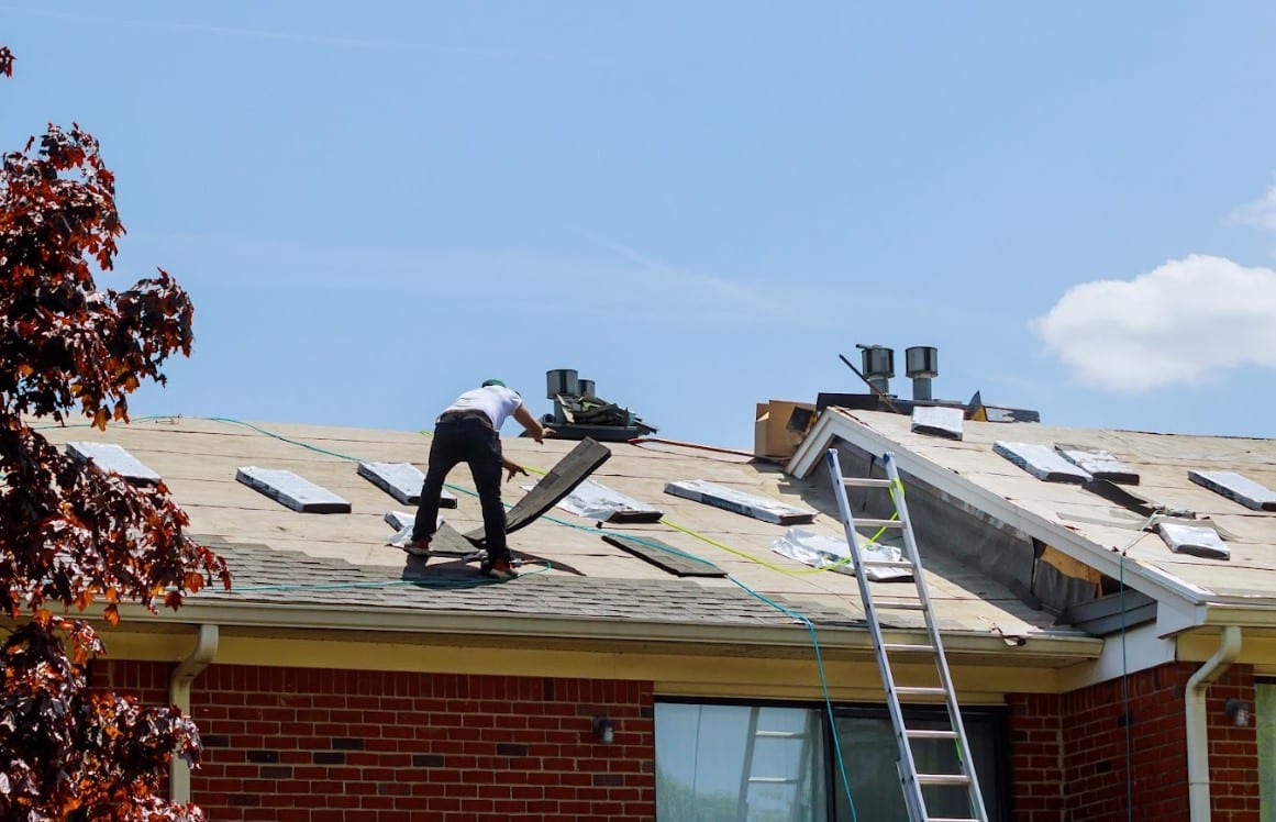 Roofer installing asphalt shingles on a residential roof with exposed decking, roofing materials, and a ladder visible.