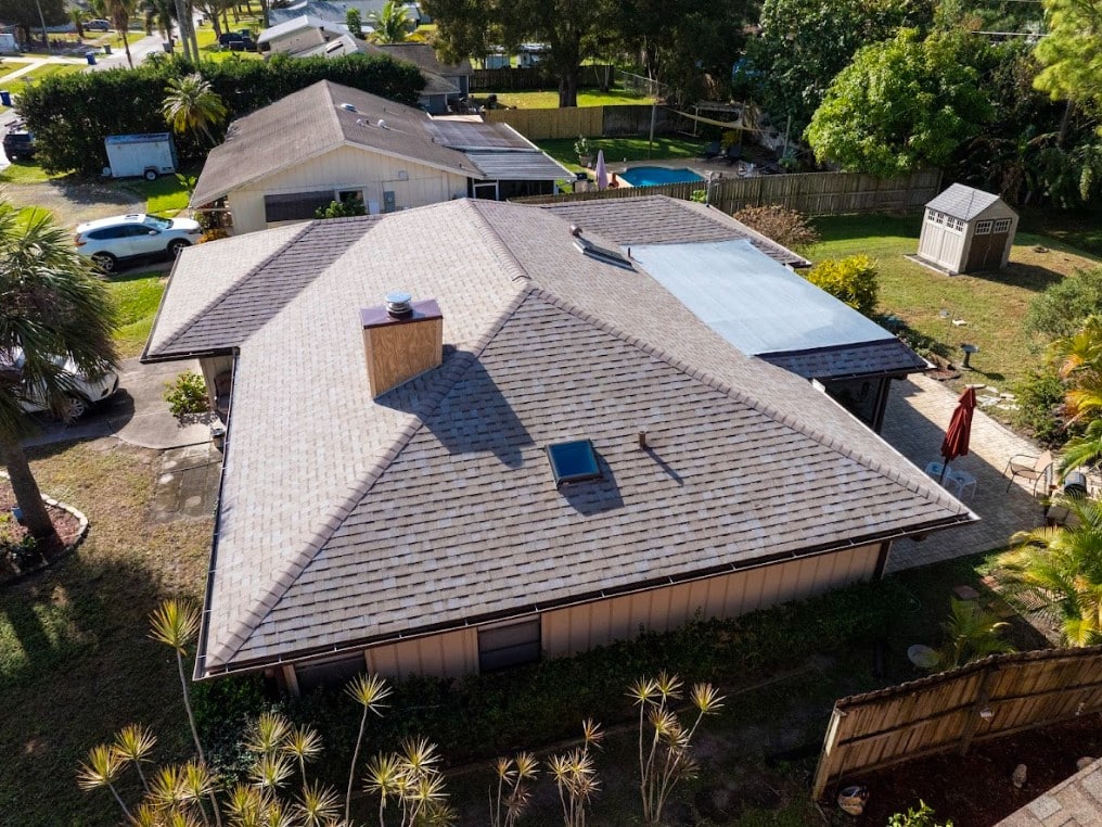 An aerial view of a single-story home's tan shingle roof with a skylight, chimney, and a small flat roof section over the patio area.