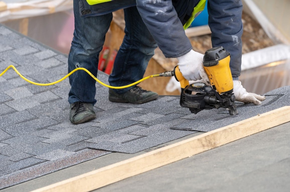 Roofer installing asphalt shingles on a residential roof while wearing a tool belt and safety equipment.
