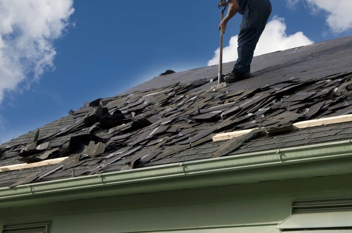 A roofer uses a tear-off shovel to strip old, damaged shingles off a house roof down to the underlayment.