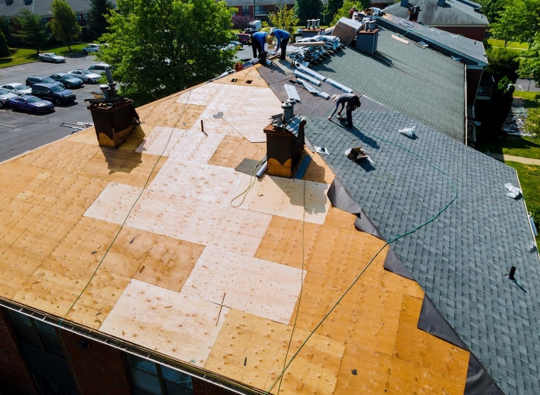 Aerial view of a roof replacement in progress with exposed wood decking, stacked shingle bundles, and roofing crews working across the roof.