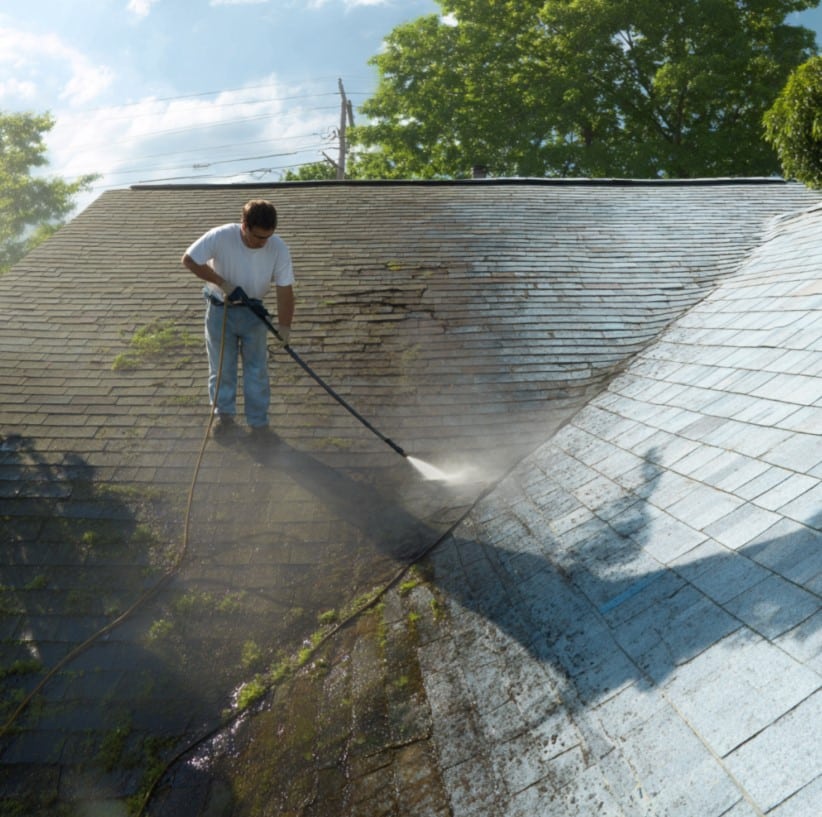 A man in a white t-shirt and jeans power-washes moss and dirt off a dirty shingle roof, revealing a clean section on the right side.