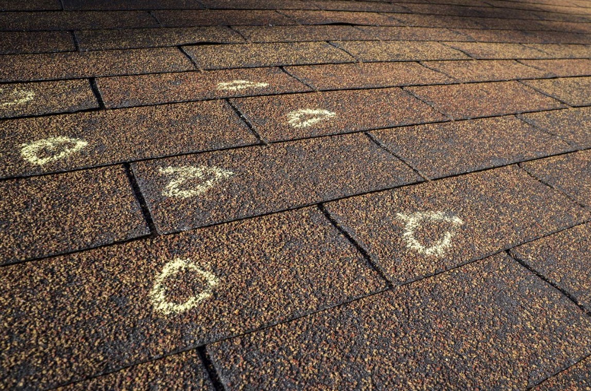 A close-up view of dark brown asphalt roof shingles with chalk circles highlighting areas of hail damage during an inspection.