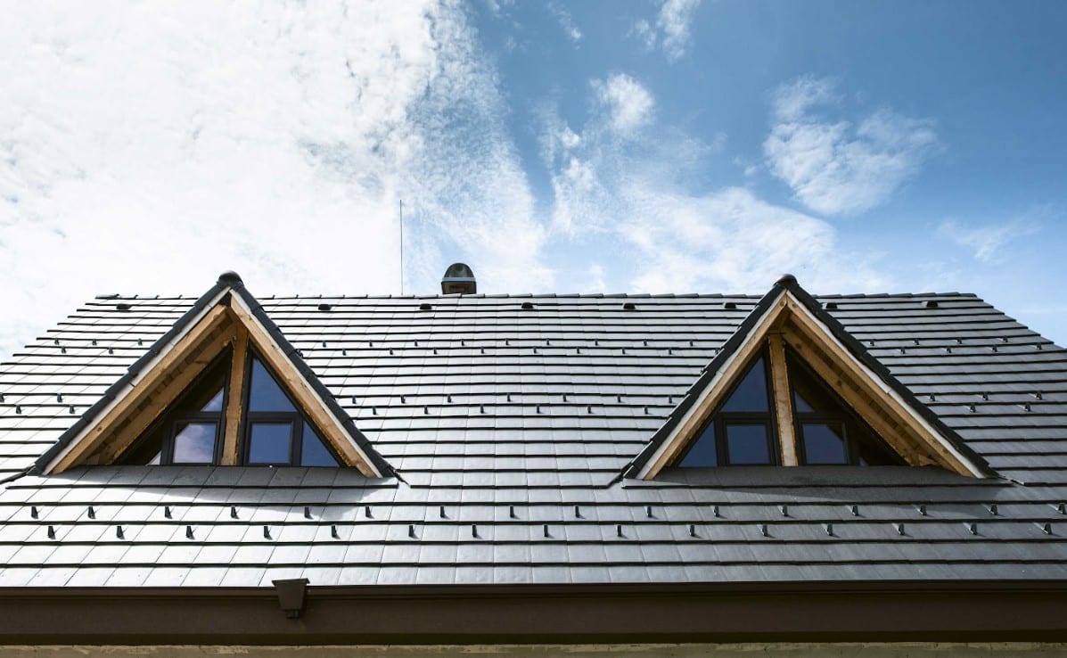 Residential home with a gray metal tile roof featuring two dormer windows and roof-mounted snow guards.