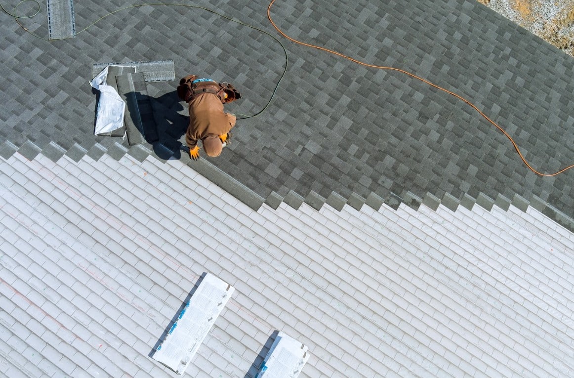 Roofer installing gray asphalt shingles on a residential roof while wearing safety equipment.