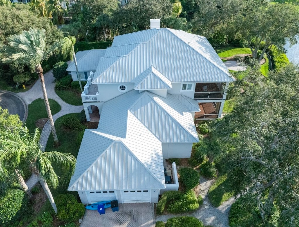 An aerial view displays a luxury waterfront home surrounded by lush tropical landscaping, featuring a light grey standing-seam metal roof with multiple hip sections and gables.