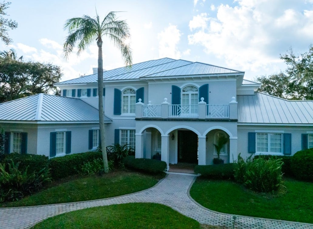 Front view of a two-story light grey coastal home with blue shutters, a paved entryway, and a light-colored standing-seam metal roof.
