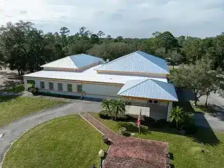 Aerial view of a large Florida building with a light metal roof, covered entrance, palm trees, and an American flag.